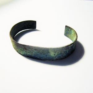 Könnte beinhalten: Ein Armreif mit verwitterter, oxidierter Oberfläche in gesprenkelten Blaugrün-, Grün- und Dunkelbrauntönen. Das Metallband hat ein schlichtes, offenes Design mit einer strukturierten, unebenen Oberfläche, die Altersspuren aufweist.