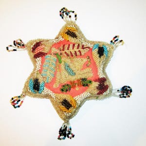 May include: A star-shaped beaded ornament with a central pink panel featuring leaf and feather designs. The star is outlined in gold beads and has colorful beaded accents and tassels at each point. A unique, handcrafted item.