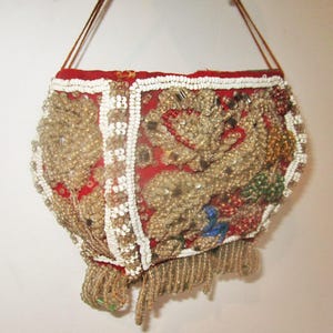 Puede incluir: Un pequeño bolso rojo de cuentas con un diseño de cuentas blancas y doradas. El bolso tiene una correa larga de color marrón y un fleco de cuentas en la parte inferior.