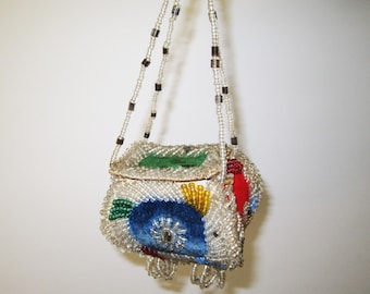 May include: A small, beaded purse with a rectangular shape and a long beaded strap. The purse is decorated with colorful beads in shades of red, blue, green, and yellow. The purse has a velvet lining and a small, round, metal clasp.