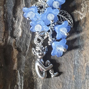 May include: A silver charm with a seated fairy, blue flower cluster, and leaf details. The charm is attached to a silver chain. The blue flowers have clear bead centres. The background is a dark, textured surface.