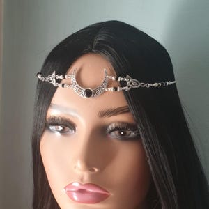 May include: Silver metal headband with a crescent moon design and a black gemstone. The headband is decorated with black beads and Celtic knot details, giving it an elegant and mystical look. The accessory is shown on a mannequin head with black hair.