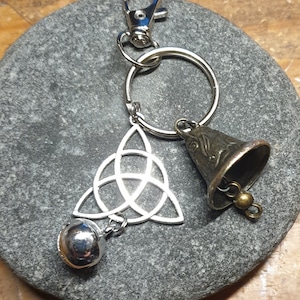May include: Silver key chain with a silver Celtic knot charm and two silver bells. The key chain is on a grey stone background.