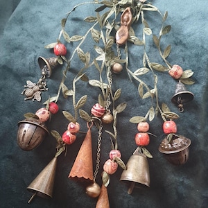 May include: Decorative hanging ornament featuring metal bells, acorn-shaped charms, and stylized leaves. The design includes red and white beaded accents, a bee charm, and a central pendant with a female figure.