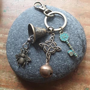 May include: A metal keyring featuring a bronze-toned lobster clasp and ring. Attached are four charms: a small bell, a detailed bee, a symmetrical Celtic knot design, and a key with a turquoise-coloured finish and a small round bell at the base.