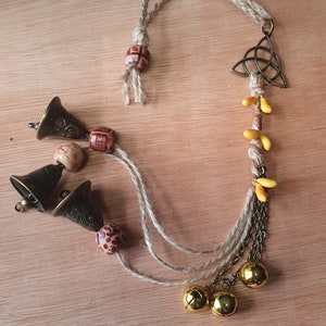 May include: A brown and gold charm with a Celtic knot design, hanging from a braided rope with yellow and brown beads. The charm is attached to three small bells and a chain with three gold bells.