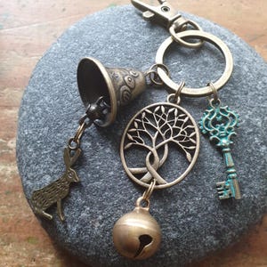 May include: A bronze-toned keyring featuring a metal ring and lobster clasp. Attached charms include a small bell, a rabbit silhouette, a circular tree of life pendant, a round jingle bell, and a key with a turquoise-coloured patina finish.