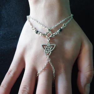 May include: Silver chain bracelet with a Celtic knot pendant and black beads. The bracelet is designed to be worn on the wrist and has a chain that extends to the finger.