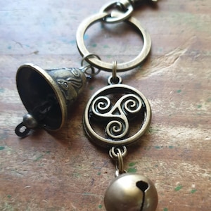 May include: Antique brass-coloured keychain featuring a bell, a round charm with a Celtic spiral design, and a small bell-shaped charm. The keychain includes a clasp and a key ring for attaching keys or other items.