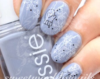 Puede incluir: Primer plano de una mano con esmalte de uñas azul claro y diseños de arte de uñas con copos de nieve y guantes blancos. El frasco de esmalte de uñas es visible en el fondo.