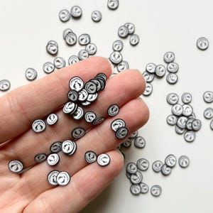 Peut inclure: Petites perles rondes en pâte polymère noire et blanche, chacune ornée d'un visage souriant. Les perles sont dispersées et tenues dans une main, probablement pour l'artisanat ou la bijouterie. Le design rappelle un personnage populaire.