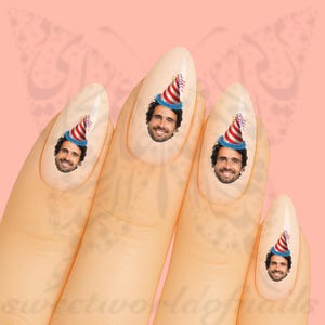 Könnte beinhalten: Nageldesign mit einem lächelnden Gesicht, das einen Partyhut mit rot-weißen Streifen und blauem Besatz trägt. Das Design wiederholt sich auf vier Nägeln, vor einem hellrosa Hintergrund mit einem Schmetterlingsdesign.
