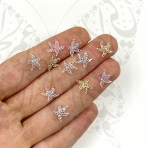 Puede incluir: Una colección de pequeños adornos en forma de estrella. Las estrellas son de varios colores, incluyendo dorado, rosa y plateado, y están cubiertas de brillantes pedrería. Estos elementos decorativos son ideales para manualidades y joyería.