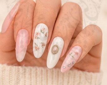Puede incluir: Una mano con uñas largas y ovaladas. Las uñas están pintadas con varios diseños, incluyendo un rosa veteado, blanco con calcomanías florales y blanco con una gema dorada. Algunas uñas tienen pequeños adornos plateados.