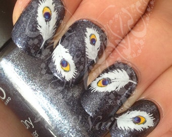 May include: Close-up of fingernails with a dark gray glitter polish and white peacock feather nail art decals. The decals feature a white feather with a blue and yellow eye.