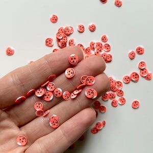 May include: Small, orange and white pumpkin-shaped confetti scattered on a white surface. The confetti has a jack-o'-lantern face design. The confetti is held in a hand. Perfect for Halloween crafts and decorations.