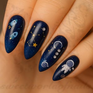 May include: Dark blue nail art with space-themed designs. Each nail features a different design, including rockets, constellations, moons, and an astronaut. The nails are almond-shaped and have a glossy finish.