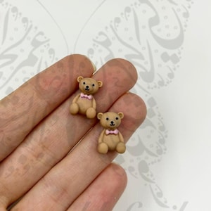 Puede incluir: Dos pequeñas figuras de osos de peluche de color canela con ojos negros y pajaritas rosas. Los osos están sentados erguidos, con los brazos a los lados. Los osos están hechos de un material liso.