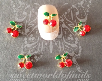 May include: Five red and green cherry nail charms with gold accents. The charms are arranged on a gray surface.