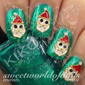 May include: Close-up of a hand with green glitter nail polish and Santa Claus nail art. Each nail features a Santa Claus face with a red hat, white beard, and gold trim. The nail polish bottle is visible.