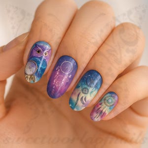 May include: A hand displaying painted fingernails with a fantasy theme. The nails are decorated with an owl, dream catchers, and celestial designs. The colour scheme includes blues, purples, and gold, giving a whimsical and artistic appearance.