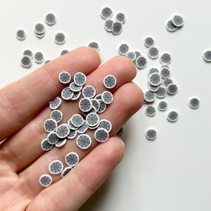 May include: A collection of small, round, white polymer clay pieces with a black spiderweb design. The pieces are scattered on a white surface and held in a hand, ideal for crafting or jewelry making.