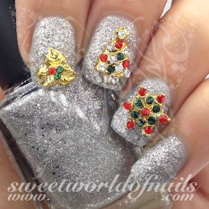 May include: Glittery silver nail polish with Christmas-themed nail art. The nails feature gold, red, green, and clear gem embellishments in the shape of a Christmas tree, holly, and a snowflake. The text "sweetworldofnails.com" is visible.