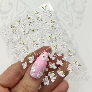 May include: White 3D flower nail stickers on a clear sheet. The stickers are arranged in a grid pattern and feature a variety of flower shapes and sizes. The stickers are perfect for adding a touch of elegance to your nails.