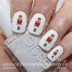 Può includere: Decalcomanie per unghie con un design a forma di schiaccianoci rosso e dorato. Le decalcomanie sono mostrate su unghie dipinte di bianco.