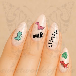Puede incluir: Una mano con uñas pintadas con diseños de uñas con temática de dinosaurios. Los diseños incluyen un dinosaurio verde azulado, un dinosaurio rosa con la palabra "ROAR", huellas de dinosaurios negras y un dinosaurio rojo con una hoja verde.