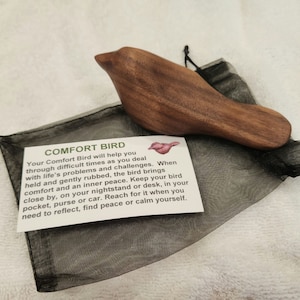 May include: A wooden comfort bird in a bird shape, resting on a black mesh bag. The bird is made of brown wood. A white card with the text "COMFORT BIRD" and a description is next to the bird.