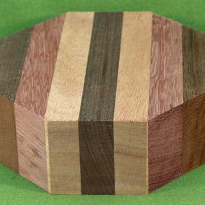 May include: A multi-colored wooden block with an octagonal shape. The block is composed of vertical strips of various wood tones, including light brown, dark brown, and reddish-brown. The wood grain is visible.