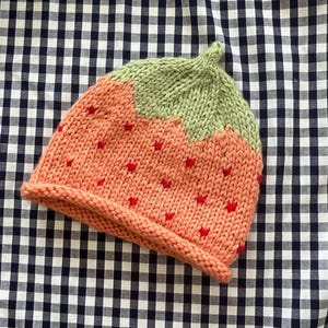 May include: A knitted hat in the shape of a strawberry. The hat is primarily peach-colored with small red heart-shaped details. The top of the hat is green, resembling the strawberry's leaves.