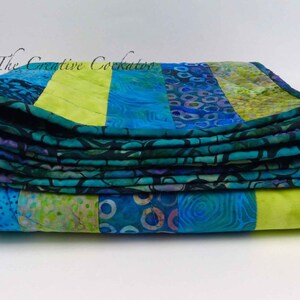 May include: A stack of folded quilts with a vibrant patchwork design. The quilts feature a combination of blue, teal, and lime green fabrics. The text "The Creative Cockatoo" is visible in the upper left corner.