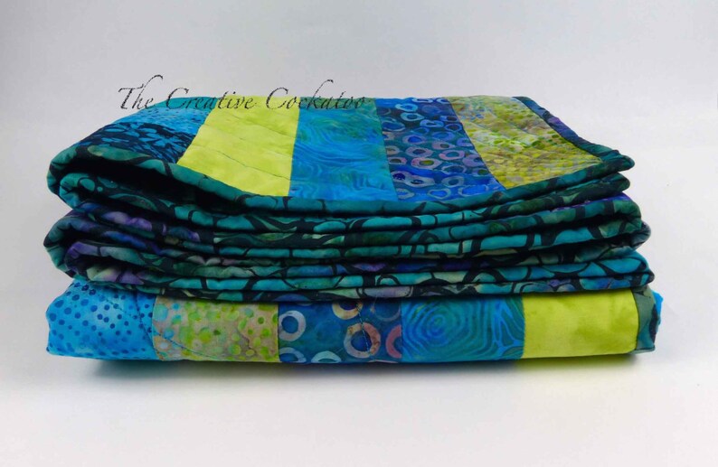 May include: A stack of folded quilts with a vibrant patchwork design. The quilts feature a combination of blue, teal, and lime green fabrics. The text "The Creative Cockatoo" is visible in the upper left corner.