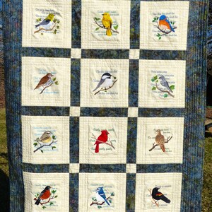 May include: A quilted wall hanging featuring embroidered birds on branches. Each bird is depicted in a square, with text above and below. The background is a patchwork of cream squares and a blue patterned fabric.