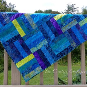 May include: A handmade quilt with a vibrant, geometric design. The quilt features a patchwork of blue, teal, purple, and chartreuse fabric rectangles. The quilt is draped over a wooden railing, showcasing its colors and textures. The Creative Cockatoo is printed on the quilt.