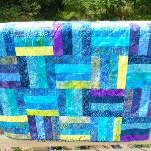 May include: A handmade quilt with a vibrant patchwork design. The quilt features rectangular blocks in shades of blue, turquoise, purple, and yellow. The fabric has various patterns, including swirls and floral prints. The quilt is displayed on a wooden railing.