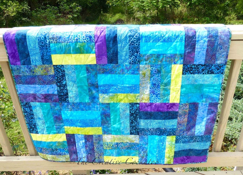 May include: A handmade quilt with a vibrant patchwork design. The quilt features rectangular blocks in shades of blue, turquoise, purple, and yellow. The fabric has various patterns, including swirls and floral prints. The quilt is displayed on a wooden railing.
