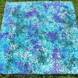 May include: A square quilt with a vibrant, abstract pattern in shades of teal, turquoise, and purple. The design features a repeating motif of interconnected, oval shapes. The quilt is displayed on a bed of green grass.