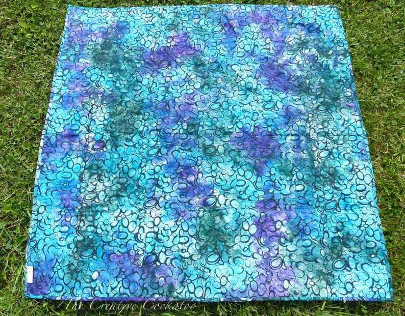 May include: A square quilt with a vibrant, abstract pattern in shades of teal, turquoise, and purple. The design features a repeating motif of interconnected, oval shapes. The quilt is displayed on a bed of green grass.