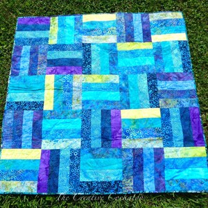 May include: A handmade quilt with a patchwork design. The quilt features rectangular blocks in shades of blue, turquoise, purple, and yellow. The quilt is made of fabric with various patterns and textures, creating a visually appealing textile.