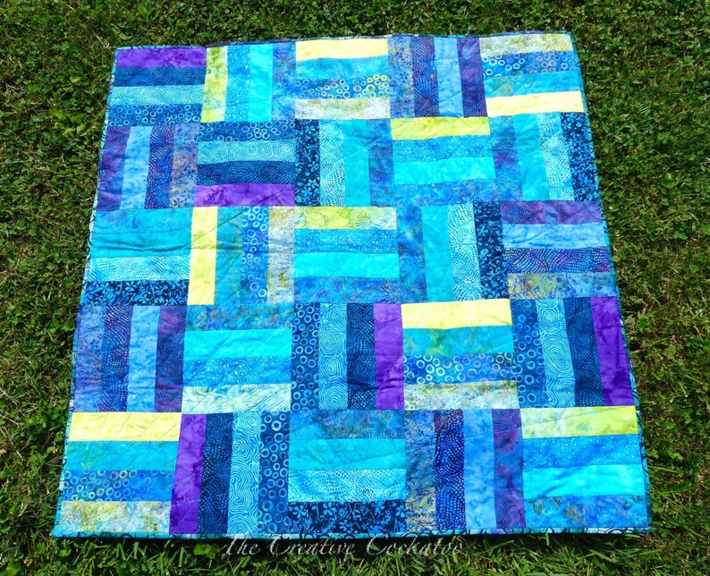 May include: A handmade quilt with a patchwork design. The quilt features rectangular blocks in shades of blue, turquoise, purple, and yellow. The quilt is made of fabric with various patterns and textures, creating a visually appealing textile.