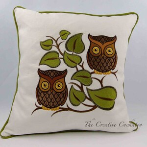May include: A white pillow with a green border features an embroidered design of three brown owls perched on a branch with green leaves. The text "The Creative Cockatoo" is embroidered in the bottom right corner.