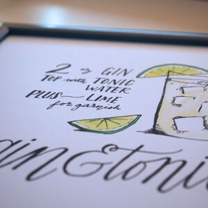 May include: A framed art print featuring a hand-drawn gin and tonic recipe. The illustration includes a glass with ice, a lime wedge, and the recipe text: "2 oz gin, top with tonic water, plus lime for garnish."
