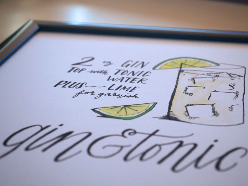 May include: A framed art print featuring a hand-drawn gin and tonic recipe. The illustration includes a glass with ice, a lime wedge, and the recipe text: "2 oz gin, top with tonic water, plus lime for garnish."