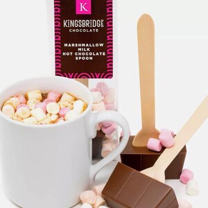 May include: A white mug filled with hot chocolate and mini marshmallows. Two wooden spoons with chocolate attached to the end. The chocolate is broken into squares. The packaging for the hot chocolate is in the background and says "Kingsbridge Chocolate Marshmallow Milk Hot Chocolate Spoon".
