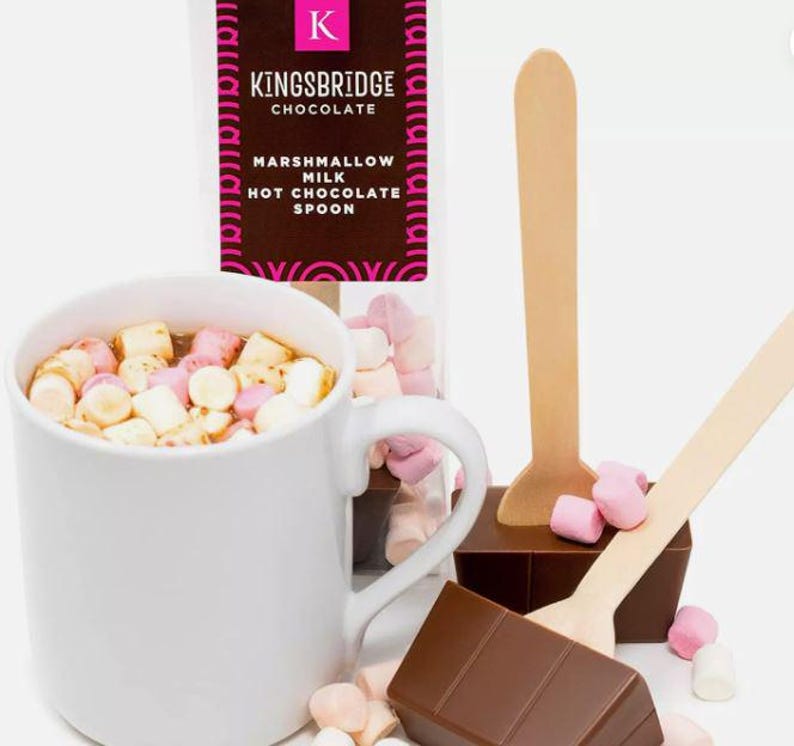 May include: A white mug filled with hot chocolate and mini marshmallows. Two wooden spoons with chocolate attached to the end. The chocolate is broken into squares. The packaging for the hot chocolate is in the background and says "Kingsbridge Chocolate Marshmallow Milk Hot Chocolate Spoon".