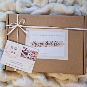 May include: A brown cardboard box with a white ribbon tied around it. The box has a label that says "A Little Something For You Hygge Gift Box" and a smaller card that says "With Love" and "To my wonderful Mum, Have a fabulous Birthday".