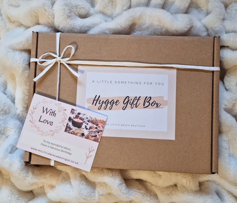 May include: A brown cardboard box with a white ribbon tied around it. The box has a label that says "A Little Something For You Hygge Gift Box" and a smaller card that says "With Love" and "To my wonderful Mum, Have a fabulous Birthday".